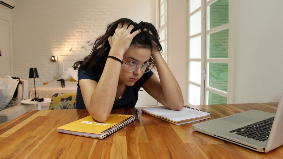 Menina de, aproximadamente, 15 anos, em seu quarto tentando estudar em uma mesa de madeira com os seus cadernos e notebook. Ela está frustrada por não estar entendendo o conteúdo da matéria.