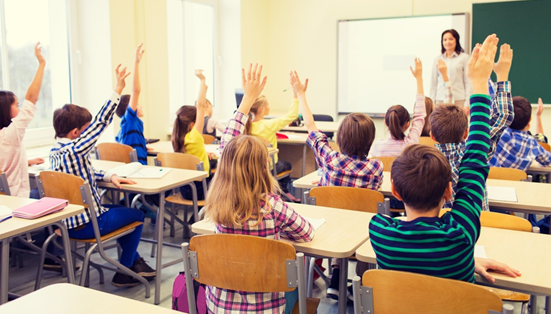 Sala de aula com diversas cadeiras e mesas marrons. Nela estão presentes vários alunos (aparentemente do Ensino Fundamental) querendo a atenção de uma única professora.