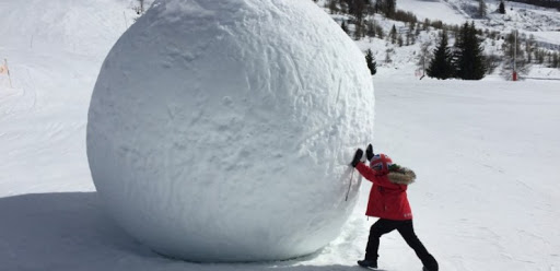 Criança agasalhada com uma calça preta, casaco vermelho e uma touca de frio vermelha, branca e azul. Ele está empurrando uma bola de neve gigante em um local que faz muito frio. O intuito dessa imagem é representar o Efeito Bola de Neve da matéria acumulada.
