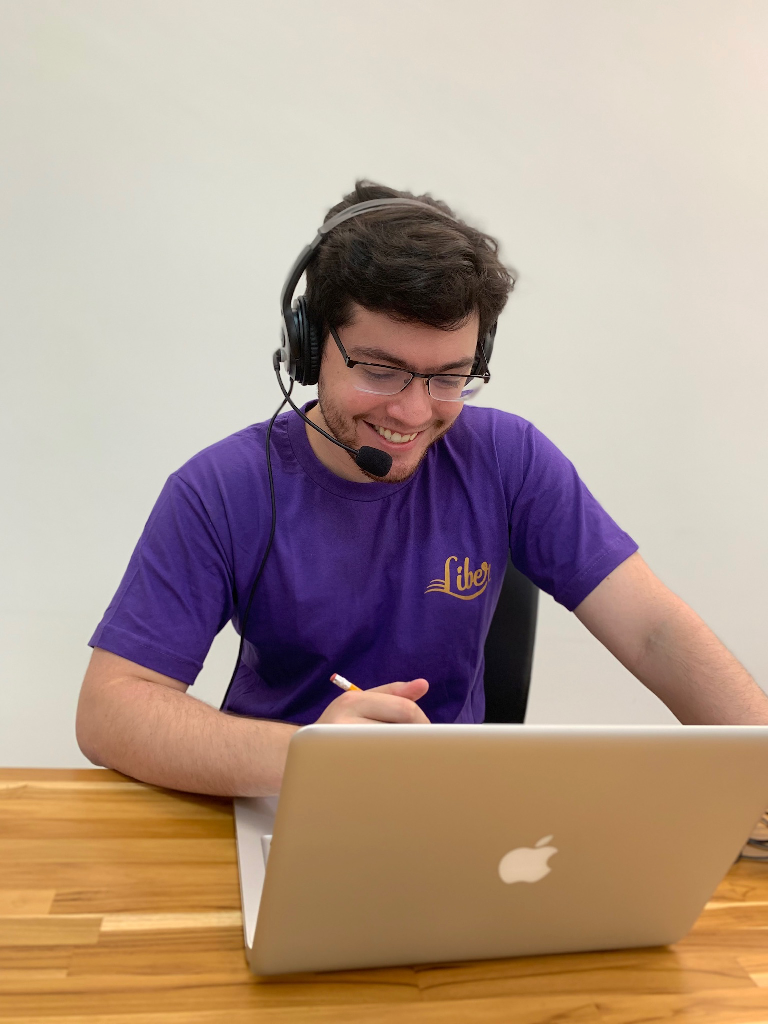 Tutor vestindo uma blusa roxa, segurando um lápis e usado um fone de ouvido com microfone. Ele está dando uma aula online com seu notebook que está apoiado em uma mesa de madeira.