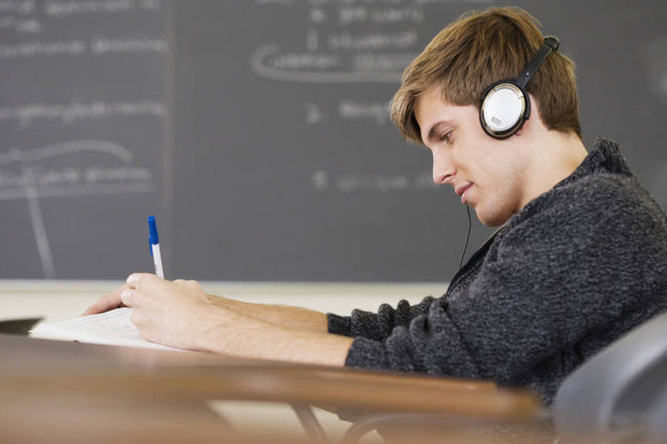 Jovem estudante de, aproximadamente, 20 anos, vestindo um casaco cinza. Ele está em um ambiente de estudos fazendo anotações em seu caderno e escutando algum conteúdo através do fone de ouvido que está utilizando, visto que o método de estudo pelo qual ele mais aprende é o auditivo.
