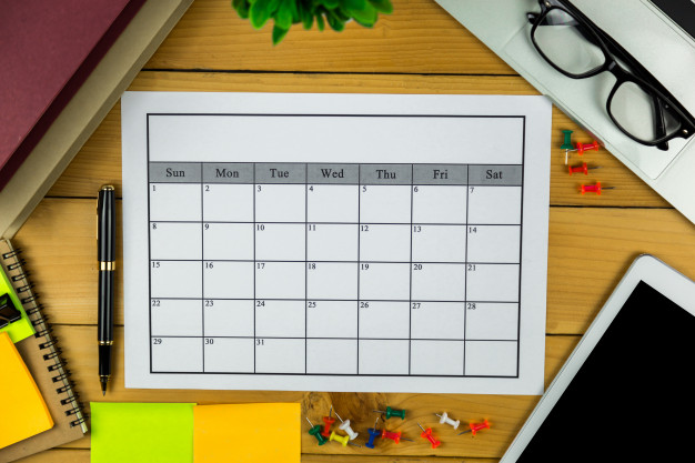 Mesa de madeira com diversos objetos em sua superfície. Calendário para ser organizado e preenchido, blocos adesivos, tablet, cadernos, , alfinetes, óculos, cadernos e caneta.