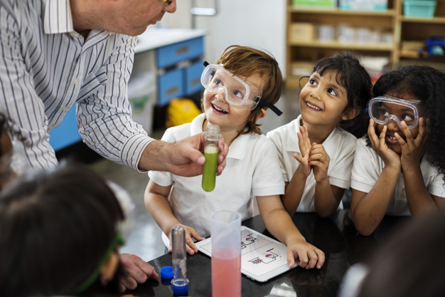 Turma de estudantes de aproximadamente 9 anos animados com uma aula em um laboratório de química com tubos de ensaio com substâncias dentro, tablet e outros materiais. Estão no ambiente um aluno, duas alunas, outros 3 alunos de costas e o professor. Eles estão testando experiências e mexendo com os objetos, esse estilo de aprendizado é o cinestésico, muito usado com crianças e jovens.